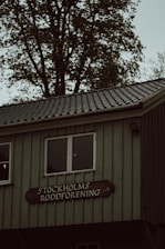 A rustic building with a wooden exterior is partially visible, featuring two square windows. Below the windows, a wooden sign reads 'STOCKHOLMS RODDFÖRENING'. The building is set against a background of tall trees with green leaves.