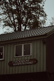 A rustic building with a wooden exterior is partially visible, featuring two square windows. Below the windows, a wooden sign reads 'STOCKHOLMS RODDF&Ouml;RENING'. The building is set against a background of tall trees with green leaves.