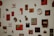 A collection of various artworks and objects mounted on a white wall. The pieces include abstract designs, photographs, and mixed media art, predominantly in red, black, and white colors. Some items are set in frames while others are standalone objects like plates or small sculptures.