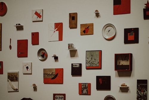 A collection of various artworks and objects mounted on a white wall. The pieces include abstract designs, photographs, and mixed media art, predominantly in red, black, and white colors. Some items are set in frames while others are standalone objects like plates or small sculptures.