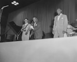 A group of musicians on stage, wearing formal suits, performing live. The setup includes a trumpet, trombone, and drums, with a piano in the background. The stage is well lit with a focus on the performers.