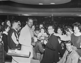 A group of people is gathered in a crowded indoor space, with a central figure holding what appears to be a piece of paper or autograph. Several individuals are reaching towards him with excited expressions. The setting suggests a busy event or gathering, and the mood is energetic.