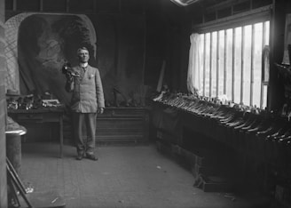 A man stands in a workshop holding a shoe in his hand. He wears a double-breasted suit and appears poised and confident. The room is filled with an array of shoes lined neatly along a table by the window, which lets in natural light. A large, artistic backdrop with a faint portrait adds depth to the space.