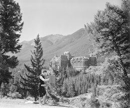 A person is painting in an outdoor setting surrounded by tall trees and mountains. In the background, a large, historic building resembling a castle is visible, set against a backdrop of forested hills.