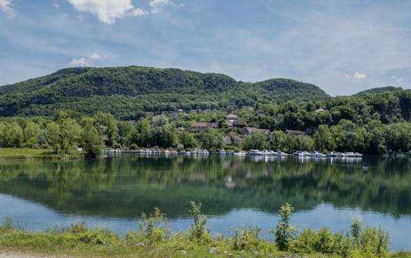 A serene lake reflects the lush green hills and trees surrounding it. In the distance, a small village with traditional buildings nestles against the hillside. Boats are moored along the shore, and a person can be seen paddleboarding on the calm water.