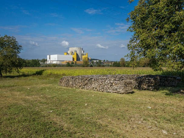 A scenic landscape featuring a grassy field with a stack of firewood in the foreground. In the background, there is a large industrial building with a mix of white and yellow colors. The sky is clear, with a few clouds, and there are trees on both sides of the image, adding to the natural setting.