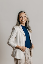 A person with long hair and a cheerful smile is dressed in a professional outfit, featuring a white blazer, a blue shirt, and beige pants with vertical stripes. The background is a plain, neutral tone.