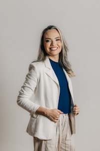A person with long hair and a cheerful smile is dressed in a professional outfit, featuring a white blazer, a blue shirt, and beige pants with vertical stripes. The background is a plain, neutral tone.