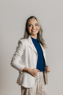 A person with long hair and a cheerful smile is dressed in a professional outfit, featuring a white blazer, a blue shirt, and beige pants with vertical stripes. The background is a plain, neutral tone.