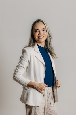 A person with long hair and a cheerful smile is dressed in a professional outfit, featuring a white blazer, a blue shirt, and beige pants with vertical stripes. The background is a plain, neutral tone.