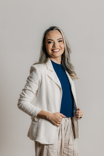 A person with long hair and a cheerful smile is dressed in a professional outfit, featuring a white blazer, a blue shirt, and beige pants with vertical stripes. The background is a plain, neutral tone.