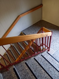 A staircase with stone steps and black anti-slip strips runs down the image. The railing is made of wood and metal, with the wood painted in a light brown and the metal in a red hue. The walls and floor are a light gray, adding a muted tone to the scene.