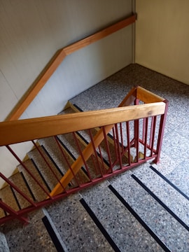 A staircase with stone steps and black anti-slip strips runs down the image. The railing is made of wood and metal, with the wood painted in a light brown and the metal in a red hue. The walls and floor are a light gray, adding a muted tone to the scene.