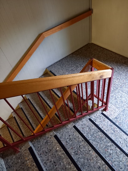 A staircase with stone steps and black anti-slip strips runs down the image. The railing is made of wood and metal, with the wood painted in a light brown and the metal in a red hue. The walls and floor are a light gray, adding a muted tone to the scene.