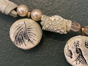A close-up view of a beaded necklace featuring large, textured beads with intricate designs. Some beads resemble pearls, while others have etched patterns, such as leaves and abstract motifs. The color palette includes muted greens and earthy tones, with metallic accents.