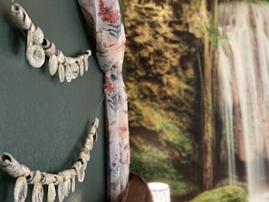 A room interior features a decorative wall with an ornamental hanging made of light-colored shapes. Next to it is a floral curtain in shades of pink and blue. In the background, a detailed mural of a lush waterfall cascades amidst rich greenery.