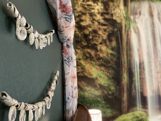 A room interior features a decorative wall with an ornamental hanging made of light-colored shapes. Next to it is a floral curtain in shades of pink and blue. In the background, a detailed mural of a lush waterfall cascades amidst rich greenery.