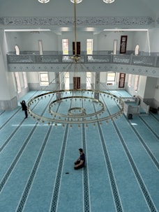 An interior view of a mosque featuring a spacious, ornately patterned turquoise carpet. A large, elegant chandelier hangs from the ceiling, adorned with gold accents and intricate designs. Tall windows with arched tops allow natural light to flood the space. Arabic calligraphy is visible on the upper wall, enhancing the serene and spiritual atmosphere. Two individuals are present: one sitting cross-legged on the carpet, appearing to be in prayer or meditation, and another standing near the entrance.