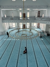 An interior view of a mosque featuring a spacious, ornately patterned turquoise carpet. A large, elegant chandelier hangs from the ceiling, adorned with gold accents and intricate designs. Tall windows with arched tops allow natural light to flood the space. Arabic calligraphy is visible on the upper wall, enhancing the serene and spiritual atmosphere. Two individuals are present: one sitting cross-legged on the carpet, appearing to be in prayer or meditation, and another standing near the entrance.