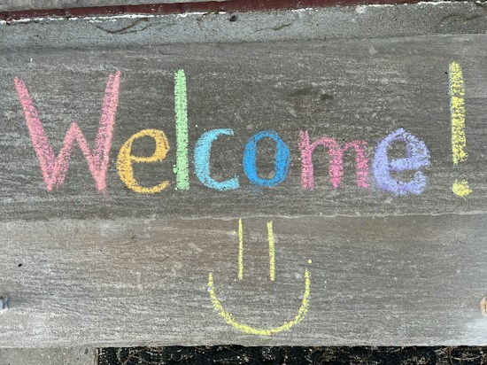The word 'Welcome!' is written in colorful chalk on a concrete surface with a smiley face drawn underneath. Each letter is in a different color, adding a vibrant and inviting feel.