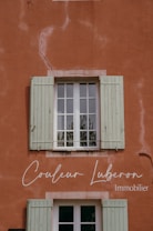 A rustic wall painted in a warm terracotta color with visible cracks and textures. A window with green wooden shutters is centrally positioned, reflecting some greenery from the outside. Elegant cursive text is displayed on the wall, indicating a business related to real estate.