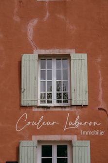 A rustic wall painted in a warm terracotta color with visible cracks and textures. A window with green wooden shutters is centrally positioned, reflecting some greenery from the outside. Elegant cursive text is displayed on the wall, indicating a business related to real estate.