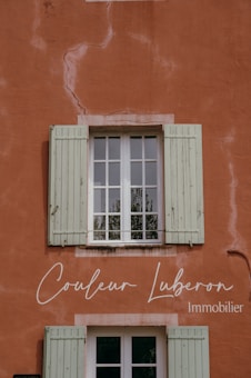 A rustic wall painted in a warm terracotta color with visible cracks and textures. A window with green wooden shutters is centrally positioned, reflecting some greenery from the outside. Elegant cursive text is displayed on the wall, indicating a business related to real estate.