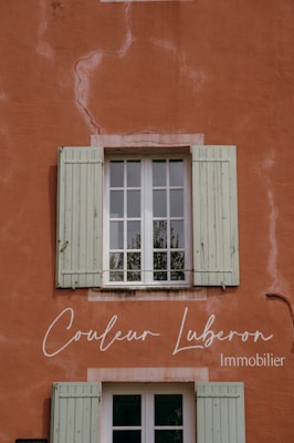 A rustic wall painted in a warm terracotta color with visible cracks and textures. A window with green wooden shutters is centrally positioned, reflecting some greenery from the outside. Elegant cursive text is displayed on the wall, indicating a business related to real estate.