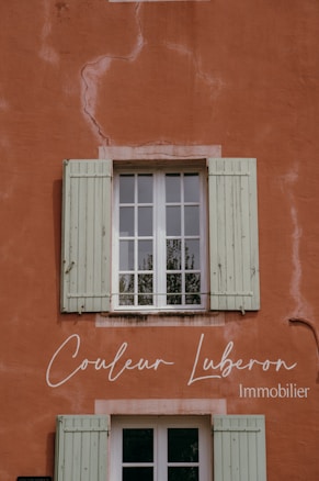 A rustic wall painted in a warm terracotta color with visible cracks and textures. A window with green wooden shutters is centrally positioned, reflecting some greenery from the outside. Elegant cursive text is displayed on the wall, indicating a business related to real estate.