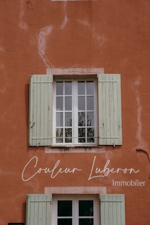 A rustic wall painted in a warm terracotta color with visible cracks and textures. A window with green wooden shutters is centrally positioned, reflecting some greenery from the outside. Elegant cursive text is displayed on the wall, indicating a business related to real estate.
