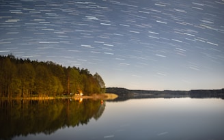 A serene night scene showing a camera on a tripod pointed at a star-filled sky with visible light trails.