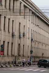 A large, beige concrete building with multiple windows in a row is situated alongside a street. Air conditioning units are mounted on the exterior walls. Several utility wires run across the scene, partially obstructing the view. Two elderly individuals are walking on the sidewalk, and a car is parked nearby. Street signs are visible along the curb.
