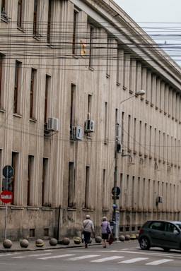 A large, beige concrete building with multiple windows in a row is situated alongside a street. Air conditioning units are mounted on the exterior walls. Several utility wires run across the scene, partially obstructing the view. Two elderly individuals are walking on the sidewalk, and a car is parked nearby. Street signs are visible along the curb.