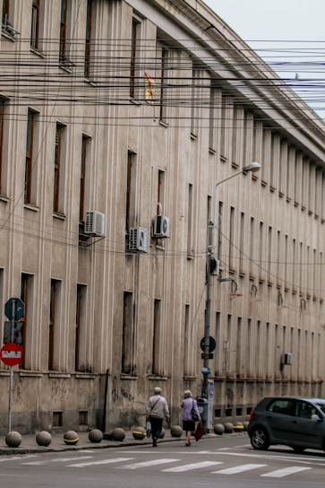 A large, beige concrete building with multiple windows in a row is situated alongside a street. Air conditioning units are mounted on the exterior walls. Several utility wires run across the scene, partially obstructing the view. Two elderly individuals are walking on the sidewalk, and a car is parked nearby. Street signs are visible along the curb.