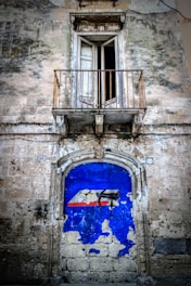 An old building facade with weathered stone walls features a large, partially peeled blue paint surface with red and white geometric shapes. Above this is a small, weathered balcony with an open window, framed by rusted metal railings.