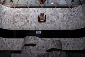 A large, wall surface covered with gray and white marble tiles features a decorative emblem with crossed keys and a crown at its center. Below, a curved staircase with matching marble is visible, with a small illuminated sign displaying 'mspl' near the bottom.
