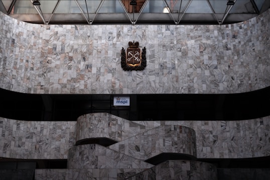 A large, wall surface covered with gray and white marble tiles features a decorative emblem with crossed keys and a crown at its center. Below, a curved staircase with matching marble is visible, with a small illuminated sign displaying 'mspl' near the bottom.