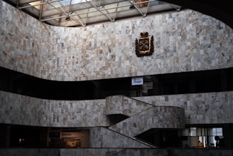 An interior view of a large building featuring marble walls with a geometric pattern. The space includes a staircase that curves upward and a metal emblem or crest mounted high on the wall. Natural light streams in from the glass ceiling, casting subtle shadows. Below are offices or display spaces with signs and modern decor.