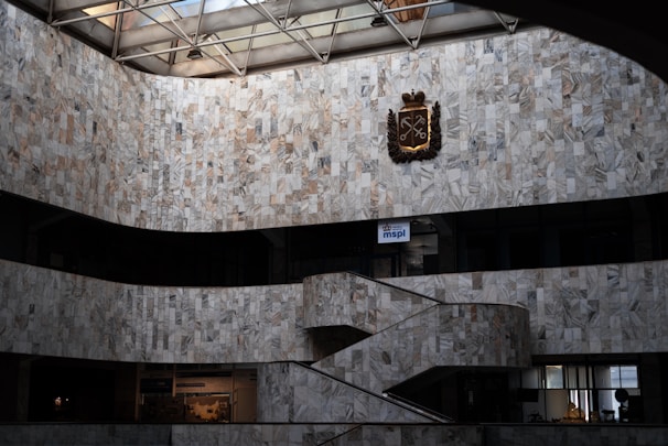 An interior view of a large building featuring marble walls with a geometric pattern. The space includes a staircase that curves upward and a metal emblem or crest mounted high on the wall. Natural light streams in from the glass ceiling, casting subtle shadows. Below are offices or display spaces with signs and modern decor.