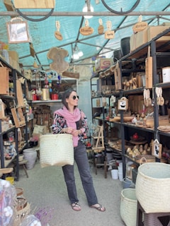 A person stands in a shop filled with wooden and woven items, holding a large woven basket. The shop has shelves lined with home decor items, baskets, and kitchenware. Wooden signs hanging from the ceiling read 'Welcome,' 'Home,' and 'Kitchen.' The person is wearing sunglasses, a pink scarf, and a floral-patterned jacket.