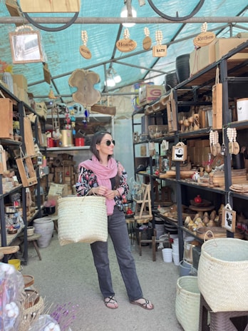 A person stands in a shop filled with wooden and woven items, holding a large woven basket. The shop has shelves lined with home decor items, baskets, and kitchenware. Wooden signs hanging from the ceiling read 'Welcome,' 'Home,' and 'Kitchen.' The person is wearing sunglasses, a pink scarf, and a floral-patterned jacket.
