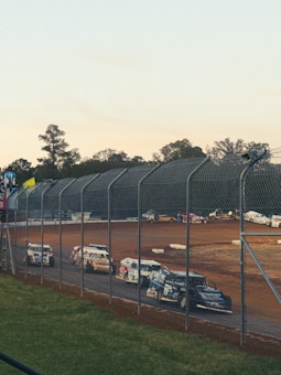 A dirt race track with several race cars lined up behind a wire fence. The cars are colorful, with numbers and advertisements on them. In the background, trees are visible under a clear sky, and the track surface is visibly reddish-brown.