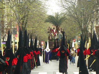 A religious procession with people dressed in traditional ceremonial robes, featuring long black pointed hoods and red sashes, marching down a tree-lined street. A large religious float with figures depicting a biblical scene is carried in the center of the procession, flanked by participants holding long staffs. The scene is set outdoors, with a background of budding trees and spectators in casual attire lining the street.