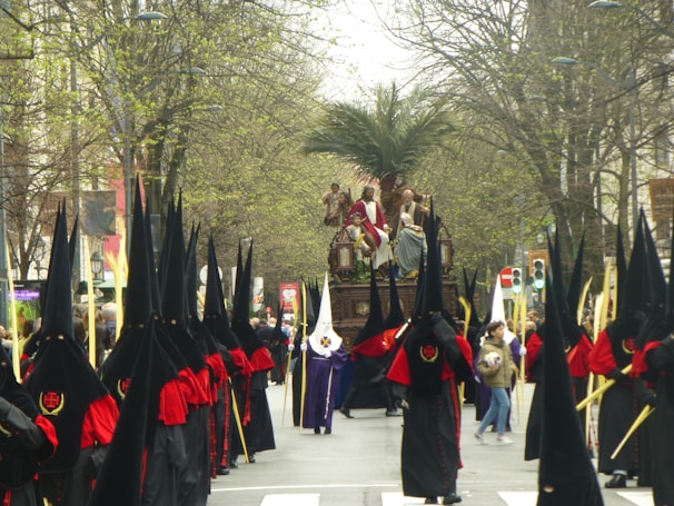 A religious procession with people dressed in traditional ceremonial robes, featuring long black pointed hoods and red sashes, marching down a tree-lined street. A large religious float with figures depicting a biblical scene is carried in the center of the procession, flanked by participants holding long staffs. The scene is set outdoors, with a background of budding trees and spectators in casual attire lining the street.