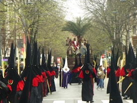 A religious procession with people dressed in traditional ceremonial robes, featuring long black pointed hoods and red sashes, marching down a tree-lined street. A large religious float with figures depicting a biblical scene is carried in the center of the procession, flanked by participants holding long staffs. The scene is set outdoors, with a background of budding trees and spectators in casual attire lining the street.