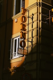 A building with a yellow exterior wall features a vertical yellow sign that reads 'FORNO' in large letters. Below this, there's a smaller sign with the word 'Pizza.' The signage casts shadows, with one part of the building in a shadow and the other illuminated by sunlight. There is also a window with a white railing and an air conditioning unit next to the sign.