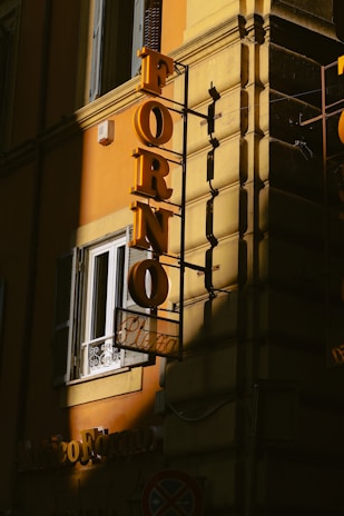 A building with a yellow exterior wall features a vertical yellow sign that reads 'FORNO' in large letters. Below this, there's a smaller sign with the word 'Pizza.' The signage casts shadows, with one part of the building in a shadow and the other illuminated by sunlight. There is also a window with a white railing and an air conditioning unit next to the sign.