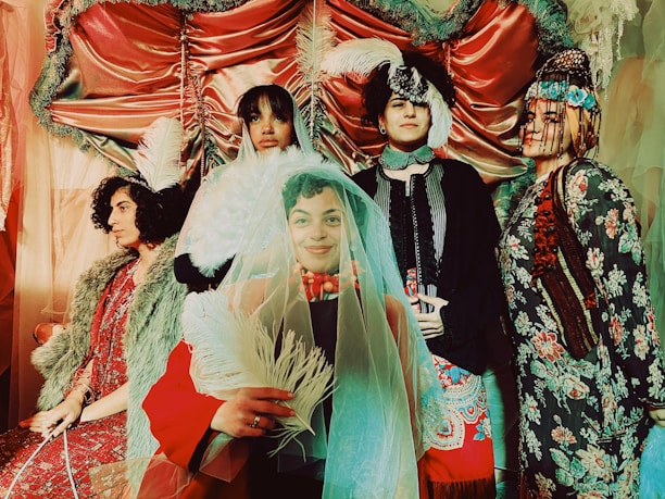 A group of five people dressed in elaborate, vintage-inspired costumes, featuring intricate patterns, fur details, and ornate headpieces. The setting is similarly dramatic, with satin drapes and soft lighting creating a theatrical ambiance. Feathers and veils add to the whimsical and eclectic style of the scene.
