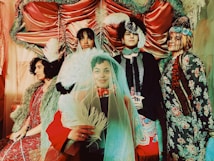 A group of five people dressed in elaborate, vintage-inspired costumes, featuring intricate patterns, fur details, and ornate headpieces. The setting is similarly dramatic, with satin drapes and soft lighting creating a theatrical ambiance. Feathers and veils add to the whimsical and eclectic style of the scene.