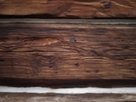 Rough-textured wooden logs with various shades from dark brown to light tan. The logs are stacked horizontally and display natural grain patterns along with some knots and minor cracks.